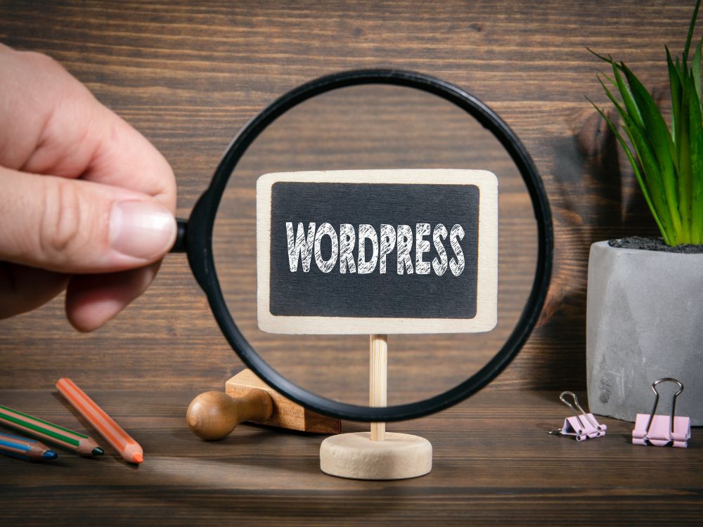 Mão segurando lupa focando na palavra WordPress escrita em placa de madeira sobre mesa com objetos, indicando que WordPress é CMS acessível.